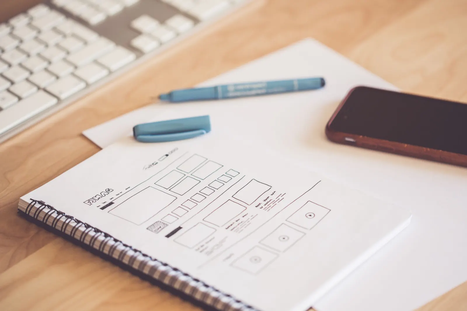 Ahşap masada, spiralli deftere çizilmiş web sayfası tel kafes (wireframe) eskizi; yanında mavi kalem, telefon ve klavye